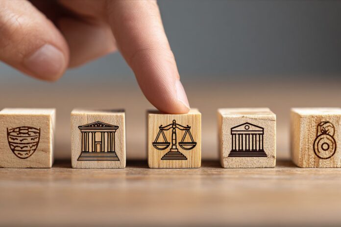 Selection of justice: Finger presses balance scale on wood block, among others symbolizing protection, courts, law enforcement, set against a simple background.